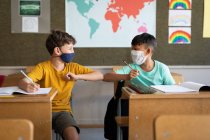 Deux garçons multi-ethniques portant des masques faciaux se saluant en touchant les coudes dans la salle de classe. Enseignement primaire distanciation sociale sécurité sanitaire pendant la pandémie de coronavirus Covid19. — Photo de stock