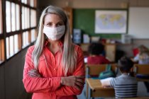 Retrato de una maestra caucásica con máscara facial de pie con los brazos cruzados en el aula. Educación primaria distanciamiento social seguridad sanitaria durante la pandemia del Coronavirus Covid19. - foto de stock