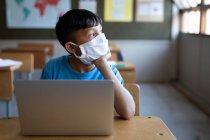 Menino de raça mista usando máscara facial sentado em sua mesa na escola, usando um computador portátil. Educação primária distanciamento social segurança sanitária durante Covid19 pandemia de coronavírus. — Fotografia de Stock