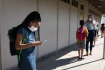 Chica de raza mixta que usa mascarilla facial usando smartphone y profesor midiendo la temperatura en la escuela primaria. Educación primaria distanciamiento social seguridad sanitaria durante la pandemia del Coronavirus Covid19. - foto de stock