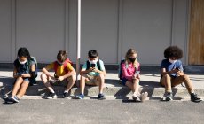 Мульти этническая группа детей начальной школы в масках для лица, используя смартфоны, сидя вместе. Начальное образование Социальное дистанцирование безопасности здоровья во время пандемии Coronavirus Covid19 — стоковое фото