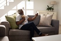 Afroamerikanischer Senior sitzt auf einer Couch, benutzt ein Smartphone, macht ein Selfie, distanziert sich sozial und isoliert sich in Quarantäne — Stockfoto