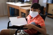 Portrait d'un garçon handicapé de race mixte portant un masque facial, assis dans son fauteuil roulant dans la classe. Enseignement primaire distanciation sociale sécurité sanitaire pendant la pandémie de coronavirus Covid19. — Photo de stock