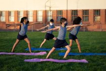 Group of multi ethnic kids wearing face masks performing yoga in the school garden. Primary education social distancing health safety during Covid19 Coronavirus pandemic. — Stock Photo