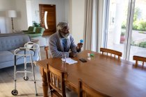 Афроамериканський старший чоловік сидить за столом у вітальні, тримаючи дві медичні пігулки і склянку води в руках, соціальну відстань і самоізоляцію в карантинному блокуванні. — стокове фото