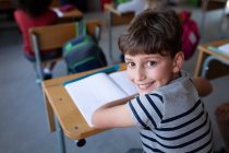 Retrato de um menino caucasiano sorrindo enquanto estava sentado em sua mesa na escola. Educação primária distanciamento social segurança sanitária durante Covid19 pandemia de coronavírus. — Fotografia de Stock