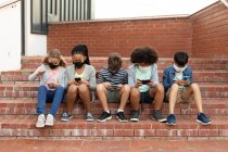 Группа мультиэтнических детей в масках для лица, сидящих на лестнице во время перерыва. Начальное образование Социальное дистанцирование безопасности здоровья во время пандемии Coronavirus Covid19. — стоковое фото