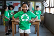 Portrait d'une fille métissée portant un t-shirt recyclé debout avec les mains sur les hanches en classe. Enseignement primaire distanciation sociale sécurité sanitaire pendant la pandémie de coronavirus Covid19. — Photo de stock