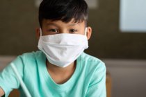 Portrait of an Asian boy sitting at desk wearing face mask in classroom. Primary education social distancing health safety during Covid19 Coronavirus pandemic. — Stock Photo