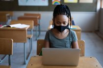Дівчина змішаної раси в масці для обличчя, використовуючи ноутбук, сидячи на своєму столі на уроці в школі. Початкова освіта соціальне дистанціювання охорона здоров'я під час пандемії коронавірусу Covid19 . — стокове фото