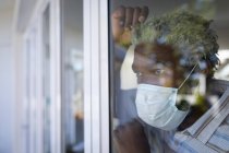 Hombre mayor afroamericano de pie en un dormitorio, con una máscara facial, mirando a través de una ventana, distanciamiento social y aislamiento en cuarentena - foto de stock