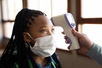Chica de raza mixta que usa mascarilla para medir su temperatura en una escuela primaria. Educación primaria distanciamiento social seguridad sanitaria durante la pandemia del Coronavirus Covid19. - foto de stock