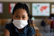 Porträt eines Mädchens mit gemischter Rasse, das mit Gesichtsmaske im Klassenzimmer sitzt und in die Kamera blickt. Grundschulbildung soziale Distanzierung der Gesundheitssicherheit während der Covid19 Coronavirus-Pandemie. — Stockfoto