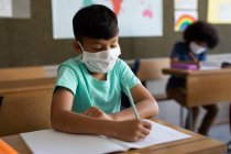 Dos niños multiétnicos sentados en escritorios con máscaras faciales en el aula. Educación primaria distanciamiento social seguridad sanitaria durante la pandemia del Coronavirus Covid19 - foto de stock