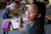 Портрет змішаної дівчини, яка посміхається, сидячи на своєму столі в школі. Початкова освіта соціальне дистанціювання охорона здоров'я під час пандемії коронавірусу Covid19 . — стокове фото