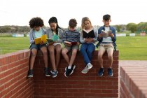 Groupe d'enfants multiethniques lisant des livres assis sur le mur pendant une pause. Enseignement primaire distanciation sociale sécurité sanitaire pendant la pandémie de coronavirus Covid19. — Photo de stock