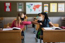 Two multi ethnic girls wearing face masks greeting each other by touching elbows in the classroom. Primary education social distancing health safety during Covid19 Coronavirus pandemic. — Stock Photo