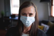 Portrait of Caucasian woman enjoying time at home, social distancing and self isolation in quarantine lockdown, wearing face mask protecting from Covid 19 coronavirus infection, looking at camera. — Stock Photo