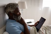 Homme âgé afro-américain allongé sur un lit dans une chambre à coucher, à l'aide d'une tablette numérique, se frottant le menton, la distance sociale et l'isolement personnel en quarantaine verrouillage — Photo de stock