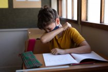 Caucasian boy sitting at desk wearing face mask in classroom, covering his face while sneezing. Primary education social distancing health safety during Covid19 Coronavirus pandemic. — Stock Photo
