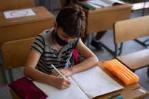 Кавказький хлопчик у масці, сидить на столі під час уроку. Первинна освіта Соціальна безпека для здоров'я під час пандемії Ковіда19 Коронавірус. — стокове фото