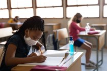Menina de raça mista usando máscara facial enquanto se senta em sua mesa na sala de aula com um desinfetante. Educação primária distanciamento social segurança sanitária durante Covid19 pandemia de coronavírus. — Fotografia de Stock