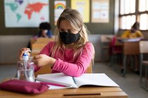 Menina caucasiana usando máscara facial enquanto sentada na mesa e higienizando as mãos. Educação primária distanciamento social segurança sanitária durante Covid19 pandemia de coronavírus. — Fotografia de Stock