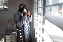 Ein afroamerikanischer Senior steht in einem Schlafzimmer, setzt eine Gesichtsmaske auf, blickt durch ein Fenster, soziale Distanzierung und Selbstisolierung in Quarantäne — Stockfoto