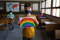Porträt eines kaukasischen Jungen mit Gesichtsmaske, der im Klassenzimmer eine Regenbogenzeichnung hält. Grundschulbildung soziale Distanzierung der Gesundheitssicherheit während der Covid19 Coronavirus-Pandemie. — Stockfoto