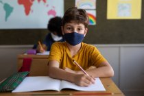 Due bambini multietnici seduti alle scrivanie con maschere facciali in classe. Istruzione primaria distanza sociale sicurezza sanitaria durante la pandemia di Covid19 Coronavirus — Foto stock