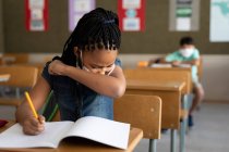 Mixed race girl sitting at desk wearing face mask in classroom, covering her face while sneezing. Primary education social distancing health safety during Covid19 Coronavirus pandemic. — Stock Photo