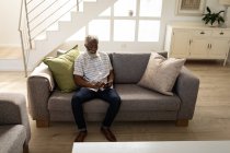 Hombre mayor afroamericano sentado en un sofá, usando un teléfono inteligente, distanciamiento social y autoaislamiento en cuarentena - foto de stock