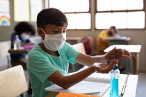 Gemischter Junge mit Gesichtsmaske, der seine Hände desinfiziert, während er auf ihrem Schreibtisch im Klassenzimmer sitzt. Grundschulbildung soziale Distanzierung der Gesundheitssicherheit während der Covid19 Coronavirus-Pandemie. — Stockfoto