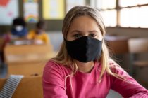Retrato de menina caucasiana sentada em mesas usando máscara facial em sala de aula. Educação primária distanciamento social segurança sanitária durante Covid19 pandemia de coronavírus. — Fotografia de Stock