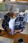 Afro-americano idoso sentado ao lado de uma mesa, limpando um smartphone com um tecido, distanciamento social e auto-isolamento em quarentena — Fotografia de Stock