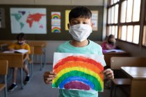 Portrait d'un garçon métis portant un masque facial tenant un dessin arc-en-ciel dans la salle de classe. Enseignement primaire distanciation sociale sécurité sanitaire pendant la pandémie de coronavirus Covid19. — Photo de stock