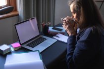 Mulher branca desfrutando de tempo em casa, distanciamento social e auto-isolamento em quarentena, sentada à mesa, usando um laptop, bebendo café. — Fotografia de Stock