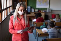 Female Caucasian teacher wearing face mask using digital tablet at school. Primary education social distancing health safety during Covid19 Coronavirus pandemic. — Stock Photo