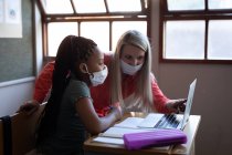Female Caucasian teacher and mixed race girl wearing face masks using laptop in class at school. Primary education social distancing health safety during Covid19 Coronavirus pandemic. — Stock Photo