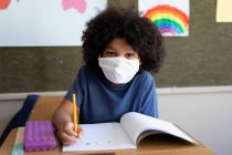 Portrait d'un garçon de race mixte assis au bureau portant un masque facial en classe. Enseignement primaire distanciation sociale sécurité sanitaire pendant la pandémie de coronavirus Covid19. — Photo de stock