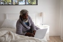 Homme âgé afro-américain allongé sur un lit dans une chambre à coucher, utilisant un ordinateur portable, distance sociale et auto isolement en quarantaine verrouillage — Photo de stock