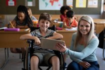 Портрет інвалідного кавказького хлопчика, що сидить на інвалідному візку і його вчительки, використовуючи табличку в класі. Первинна освіта Соціальна безпека для здоров'я під час пандемії Ковіда19 Коронавірус. — стокове фото