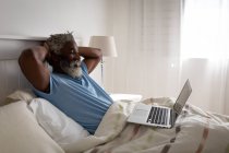 Uomo anziano afroamericano sdraiato su un letto in una camera da letto, utilizzando un computer portatile e sorridente, distanza sociale e isolamento in quarantena — Foto stock