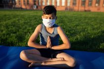 Хлопчик змішаної раси в масці для обличчя, що виконує йогу в шкільному саду. Початкова освіта соціальне дистанціювання охорона здоров'я під час пандемії коронавірусу Covid19 . — стокове фото