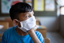 Thoughtful mixed race boy wearing face mask sitting on his desk at school. Primary education social distancing health safety during Covid19 Coronavirus pandemic. — Stock Photo
