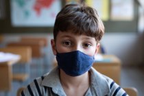 Retrato de un chico caucásico con una máscara facial, sentado en su escritorio en clase en la escuela. Educación primaria distanciamiento social seguridad sanitaria durante la pandemia del Coronavirus Covid19. - foto de stock