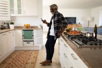 Старший афроамериканец, стоящий на кухне, используя смартфон, социальную дистанцию и самоизоляцию в карантинной изоляции — стоковое фото