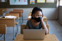 Gemischtes Mädchen mit Gesichtsmaske und Laptop, während es in der Schule auf seinem Schreibtisch sitzt. Grundschulbildung soziale Distanzierung der Gesundheitssicherheit während der Covid19 Coronavirus-Pandemie. — Stockfoto