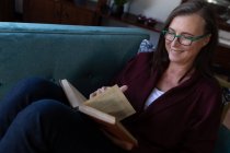 Kaukasische Frau genießt Zeit zu Hause, soziale Distanzierung und Selbstisolierung in Quarantäne, liegt auf dem Sofa im Wohnzimmer, liest ein Buch. — Stockfoto
