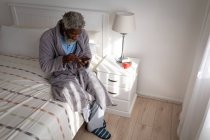 Uomo anziano afroamericano seduto su un letto in una camera da letto, utilizzando uno smartphone, distanza sociale e isolamento in quarantena — Foto stock